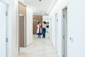 Fototapeta premium Female doctor in a white coat and scrubs walking through a modern medical clinic hallway with a young patient. Professional healthcare consultation and communication in a bright, clean hospital wing.