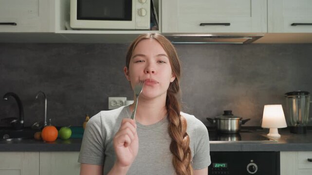 Smiling woman tasting food at counter, holding fork, braided hair, stove and microwave visible, bright lamp light, playful expression, vlogger energy, casual gray shirt, recipe sampling