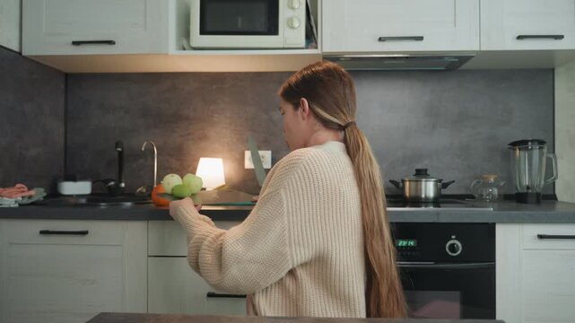 Pregnant white woman seated in kitchen wearing knit cardigan and bralette, hands resting on visible belly, soft lamp glow, apples and orange on counter, contemplative prenatal moment, slow morning,