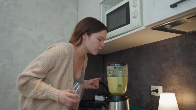 Pregnant caucasian woman blends green smoothie at kitchen countertop, holding measuring cup and glass, pouring ingredients into blender, leaning to check aroma, cozy cardigan, microwave overhead, warm