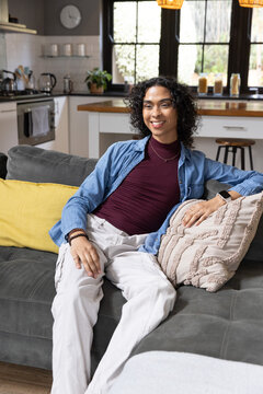 Non-binary person sitting on dark gray sofa at home wearing maroon top, denim shirt, smartwatch
