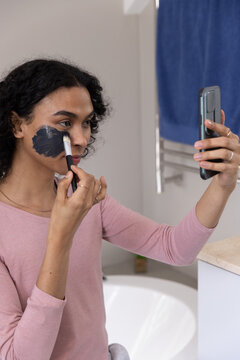 Indian non-binary applying charcoal face mask with flat brush on bathtub edge holding smartphone