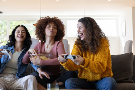 Diverse female friends sitting on sofa playing game holding white controllers and sharing chips