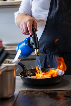 Sc&egrave;ne dynamique d'un chef en tablier noir utilisant un chalumeau de cuisine bleu pour flamber des aliments, avec des flammes vives s'&eacute;levant d'une po&ecirc;le au c&oelig;ur d'une cuisine professionnelle.