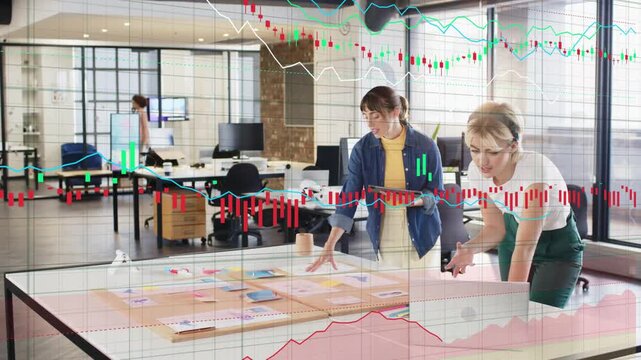 Two women planning at table tapping tablet and moving cards, finance charts overlaying for strategy