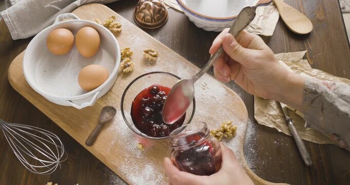 Woman hands taking cherries from jar with spoon into small glass bowl. Baker preparing sweet filling with berry jam for homemade cake on wooden board. Culinary process of making delicious dessert with
