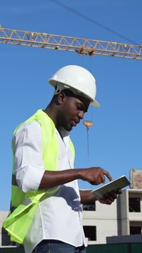 Black Inspector Using Tablet Construction Site, Wearing Hard Hat And Neon Vest, Crane And Concrete Background, Tapping Screen To Review Plans And Checklists, Bright Sunny Day, Focused Professional