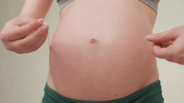 Closeup pregnant belly measured with tape, green pants visible. Hands securing soft tape around abdomen, gentle indoor lighting, calm clinical mood, monitoring trimester growth and progress, home