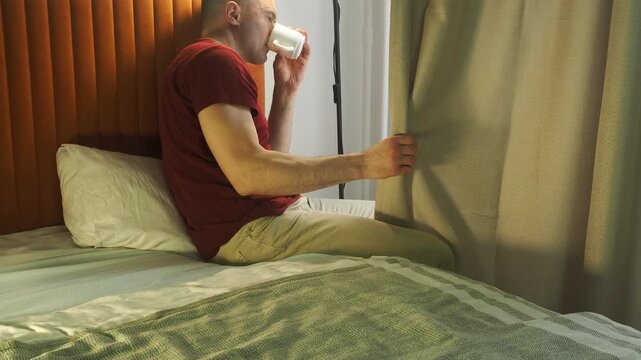 A man drinking coffee in bed looking out the window