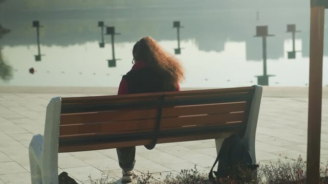 Solitary person on bench by waterfront, curly hair profile in late morning sunlight creating soft reflection on calm harbor pier posts and distant signage frame scene, bin and bag at feet, cinematic