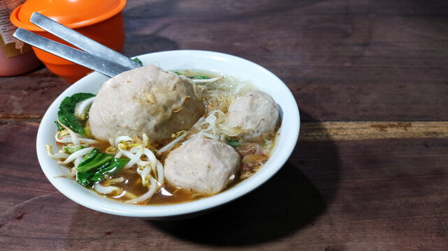 A bowl of traditional Indonesian bakso soup featuring large beef meatballs, noodles, bean sprouts, and green vegetables served in savory broth on a rustic wooden table.