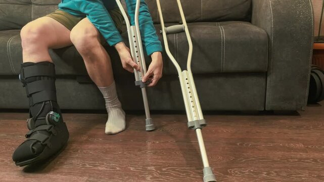 Man sitting on sofa positioning crutches for support, recovery preparation, rehabilitation and mobility concept.