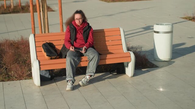 Young woman on bench looking up during quiet afternoon, red jacket and backpack beside seat, resting between errands with contemplative gaze, empty promenade and long shadows, still urban scene