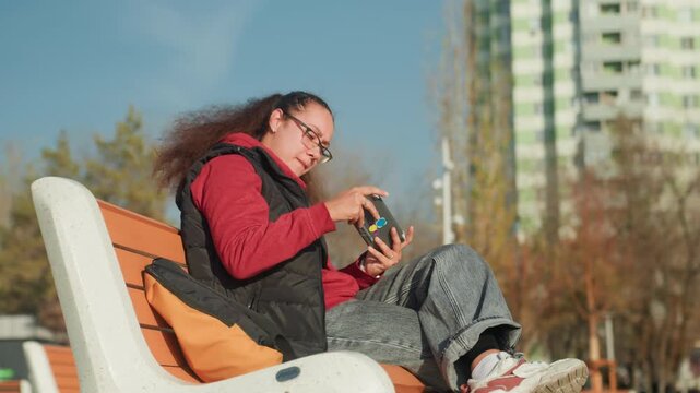 White woman focused on smartphone screen while seated on modern outdoor lounge, highrise apartment towers in background, thumbs tapping and hair moving in breeze, bright midday sunlight, active