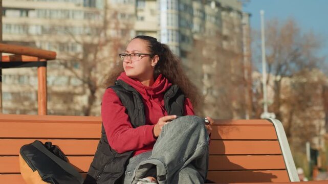 White woman sits on urban bench, leaning back to feel sunlight, wearing red hoodie and black vest, gray pants and glasses, backpack at side, scrolling phone and gazing at skyline, mood shifts