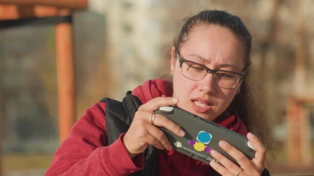 Woman playing mobile game outdoors, frustrated expression on bench under autumn sunlight, wearing glasses and red hoodie, gripping phone controller, focused gestures, candid urban park background,