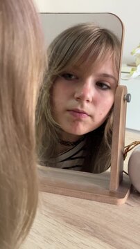 Thoughtful teenage girl closely examining her reflection in a wooden desk mirror, focusing on skin imperfections with a serious expression, reflecting on self-image and beauty standards