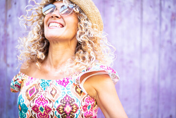 Fototapeta premium Happy woman with curly blonde hair, sunglasses, and a straw hat is feeling joy and freedom while smiling and looking up against a lavender background, embracing a carefree summer lifestyle