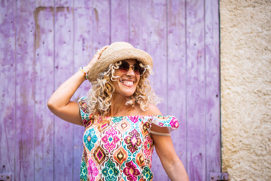Cheerful woman in a straw hat and colorful patterned dress smiling broadly against a vibrant purple wooden door, enjoying a sunny summer day outdoors, half-body frontal view