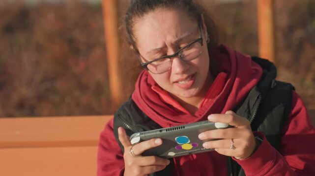 Goldenhour bench woman gaming with smartphone, red hoodie and glasses, shifting emotions from furrowed concentration to relieved smile, looking up at sunset sky, warm rim light and autumn foliage