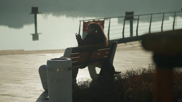 Person on bench by waterfront, hooded silhouette facing misty pier early morning mood with lifebuoy and railing, muted tones, contemplative posture suggesting commuter pausing between journeys,