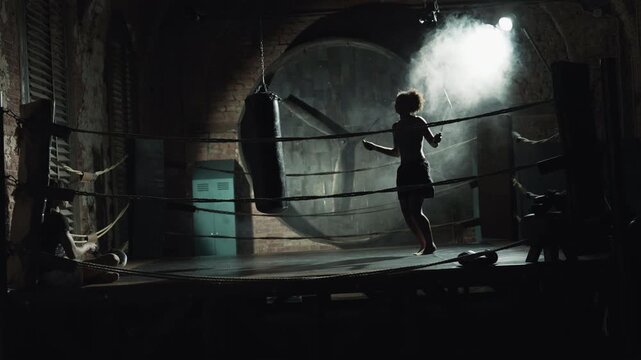 Woman skipping rope on boxing ring in dark gym, cardio workout for healthy life. Slender african american lady training alone in fitness center, future champion preparing to important competition