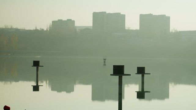 Misty urban lake with reflections, distant apartment skyline mirrored on glassy water, fogsoftened horizon, silhouetted posts and buoys punctuating still surface, soft greengrey palette, early morning