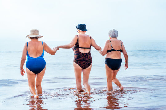 Three senior women friends holding hands, walking into the ocean water together, enjoying a refreshing summer day on their beach vacation expressing friendship and active retirement