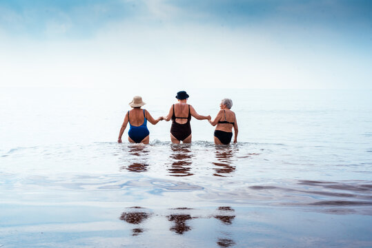 Three senior women wading into the ocean holding hands, sharing a moment of enduring friendship and active leisure during a summer vacation, reflecting companionship and wellness
