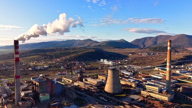 Aerial landscape of an active power station with cooling towers, generator units, and smoke against a mountain backdrop. Thermal power plant facility.