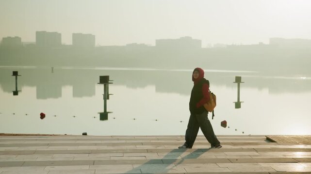 Man walking along calm waterfront at sunrise. Long shadow stretches across promenade, reflective water mirrors distant skyline, purposeful stride with backpack, misty horizon and golden light, mood