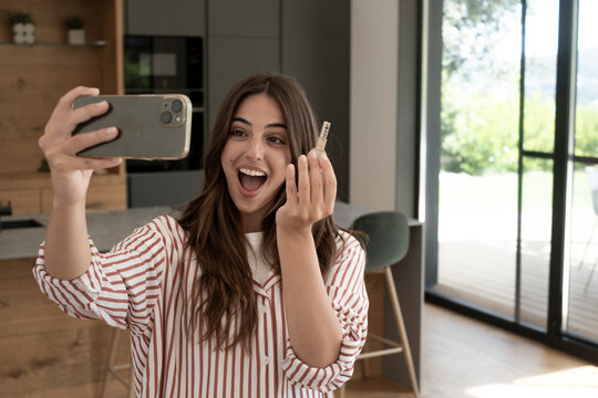 Happy young woman joyfully holding new home keys and smiling at a smartphone while recording a video or taking a selfie in her modern apartment, celebrating home ownership