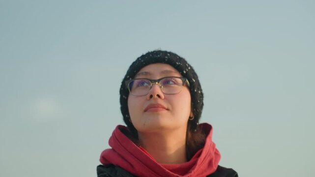 Young woman gazing at sunrise beach in beanie and glasses, red hoodie quiet introspective portrait with soft sky, distant waves, cold air and faint snow flakes on hat, student traveler taking a deep