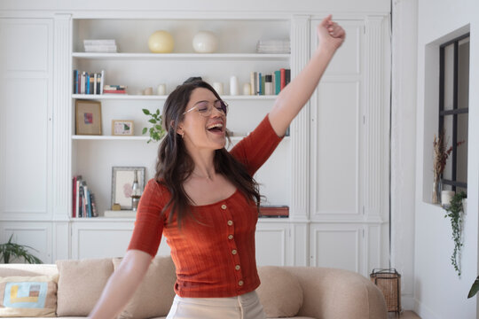 Woman feeling cheerful and expressing elation with a raised arm, enjoying a moment of happiness and victory while dancing relaxed in her modern living room