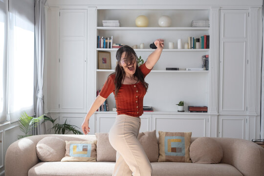 Young woman enthusiastically dancing and celebrating with arms raised in a modern living room, finding joy and freedom while listening to music at home
