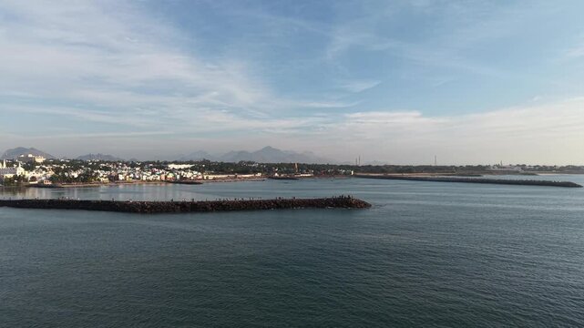 Wide coastal view showing a long rock breakwater stretching into the sea with people walking and gathering on it.