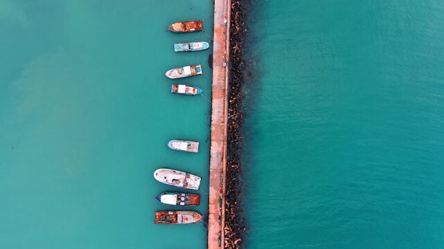 Aerial view of a concrete jetty lined with fishing boats in turquoise water.