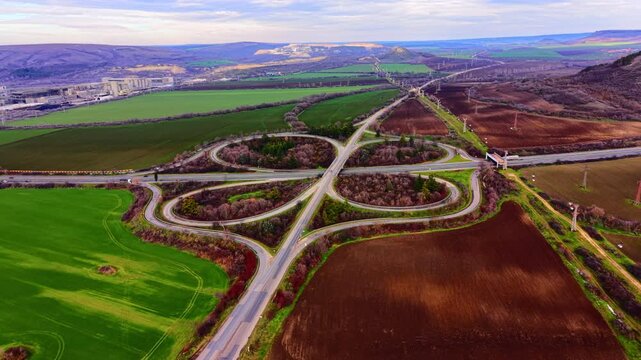 Road junction surrounded by green fields with an industrial plant in the background. Aerial view of highway cloverleaf interchange near factory.