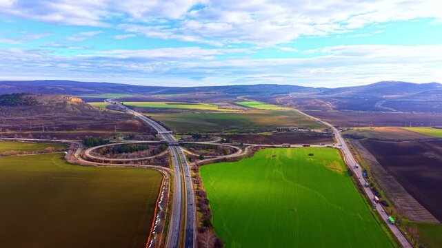 Aerial view of a complex highway cloverleaf interchange. Asphalt roads connect through green and brown agricultural fields. Highway interchange junction in a rural landscape area.