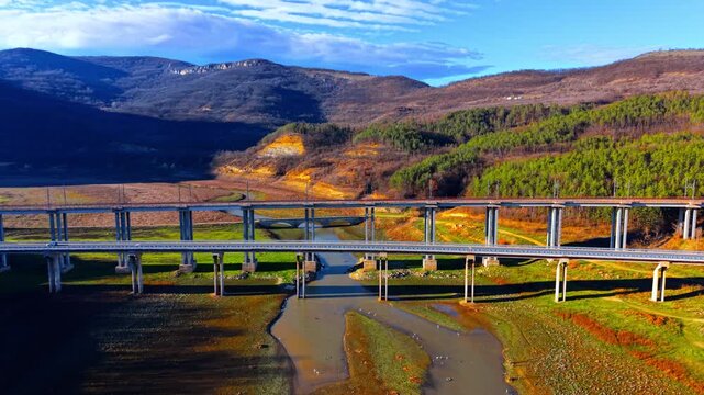 Concrete bridge pillars supporting a highway and a railway track over a drying river bed and green hills. Modern bridges and railway viaduct crossing a river valley.