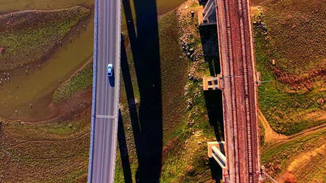 Aerial shot of a cars driving on a concrete highway bridge parallel to a red gravel railway bridge over a river valley.
