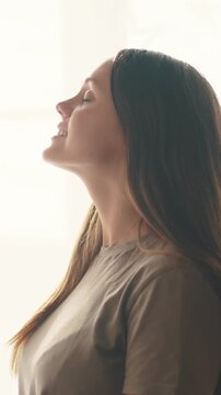A woman stands in profile, looking upwards toward the light. Her long hair falls gracefully, and her expression reflects a moment of calm and contemplation.