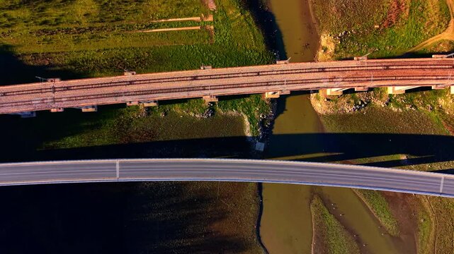 Curved asphalt highway and a straight railway bridge crossing a shallow river and green fields. Top down aerial view of parallel road and railway bridges over river.