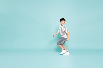 Fototapeta premium Happy young Asian boy in a striped polo shirt running cheerfully against a teal background. Childhood energy, active lifestyle, and healthy growth
