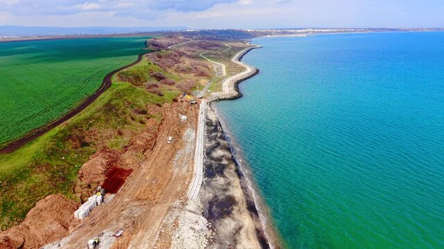 Coastline featuring sea wall construction using concrete blocks next to green fields and a turquoise sea under a cloudy sky. Aerial view of coastal fortification and road construction.