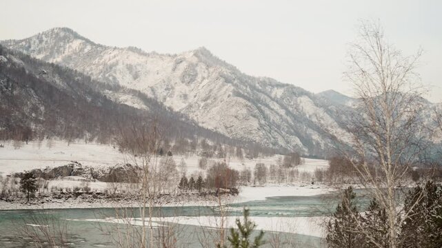 Snowy mountain river valley at dawn, birch trees along frozen shoreline, teal water with thin ice, mist rising from river, overcast soft light, remote alpine peaks covered in snow, cinematic aerial