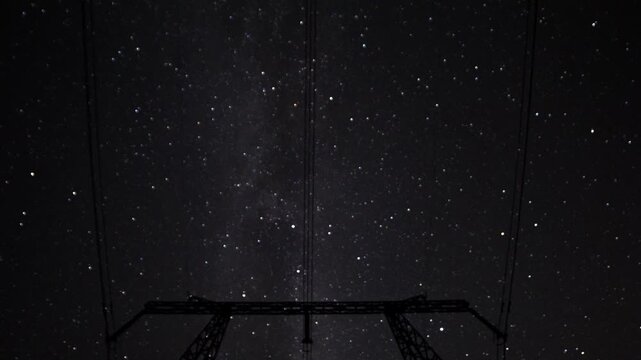 Milky Way arch above silhouetted pylon, long exposure night sky during Perseids, faint meteor streaks, powerlines crossing frame, remote field horizon, deep blue tones, calm contemplative mood