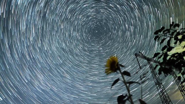 Spiral star trails framing sunflower silhouette, powerful circular motion from long exposure, cool cyan hues, textured petals and rural tower outlines, Perseids hints and hypnotic nocturnal atmosphere