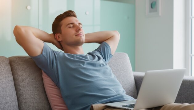 Young man relaxing on a sofa with eyes closed and hands behind head for remote work lifestyle, mental health awareness and home wellness campaigns