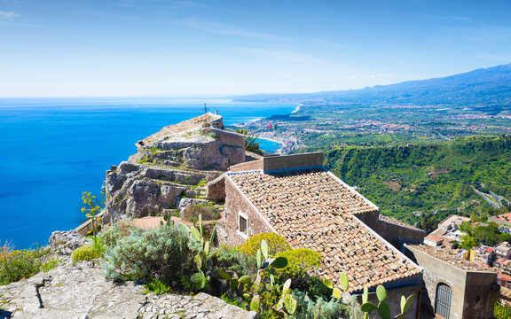 Panoramic view from Chiesa Madonna della Rocca above Taormina, Sicily. Stone chapel and tiled roof on rocky cliff overlooking Ionian Sea, coastline, town and green hills under clear blue sky